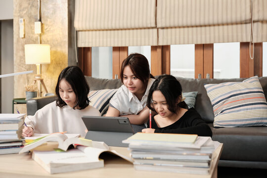 Three Asian teenage girls concentrate on online class, remote learning at home using tablet, writing and reading textbooks, self-study for back-to-school, Homeschooling education homework lifestyle.