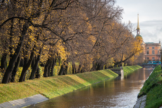 Autumn trees line narrow canal leading toward Mikhailovsky Castle in central Saint Petersburg. Golden foliage, grassy embankments and calm water surround historic urban landmark during late season.