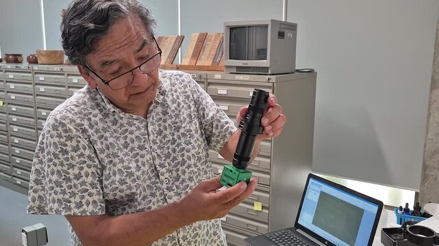 Elderly Hispanic researcher wearing glasses screws a tube onto a digital microscope camera while working in a wood collection library. Scientific research, botany and forestry concept.