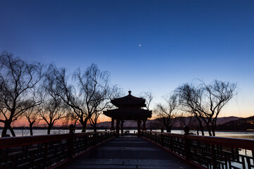 A traditional pavilion stands on a bridge over a lake at twilight, with bare willow trees under a clear sky. © tai