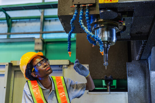 Female operator visually inspects the spindle and coolant nozzles of a CNC machine before operation. Wearing safety glasses, gloves, and a yellow hardhat.