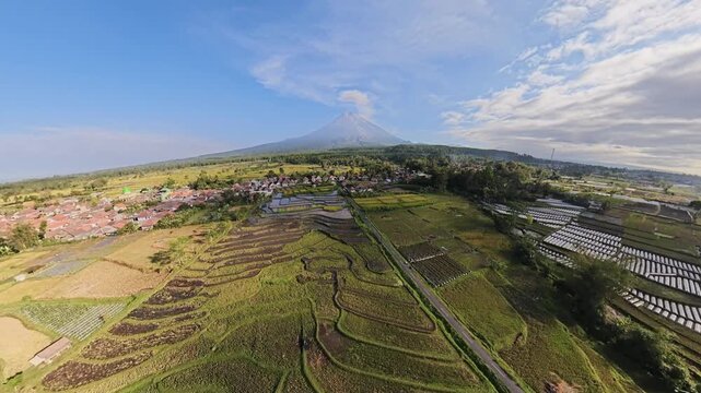 Drone fisheye view showing terraced rice paddies, village and Mount Semeru under blue sky