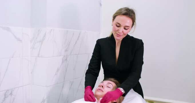 Facial cleaning. Beauty treatment. Esthetician in gloves removing mask from female client skin using cotton sponge in modern salon.
