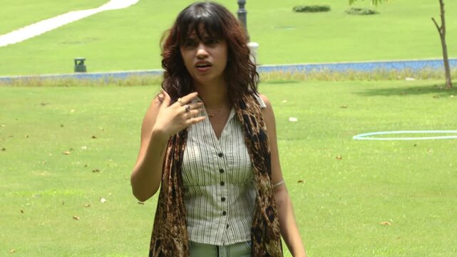 Young Indian woman experiencing discomfort and sweating while walking outdoors in green city park during extreme summer heat, heatwave, rising temperatures, climate change and global warming.