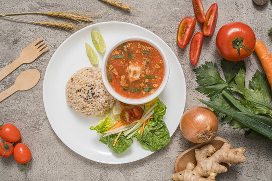 Tomato soup served in a bowl, accompanied by red rice, orange slices, and a side salad arranged on a plate, complete with fresh ingredients used as props.