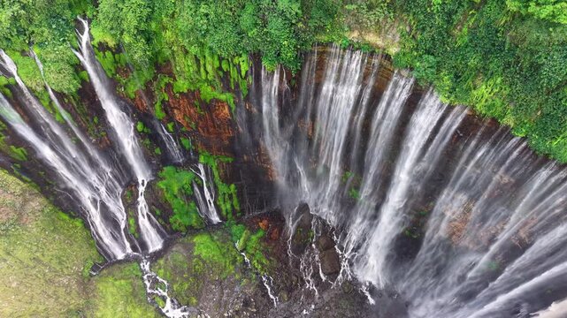 Multiple waterfalls cascade down lush green cliffs. Water rushes over mossy rocks at Tumpak Sewu