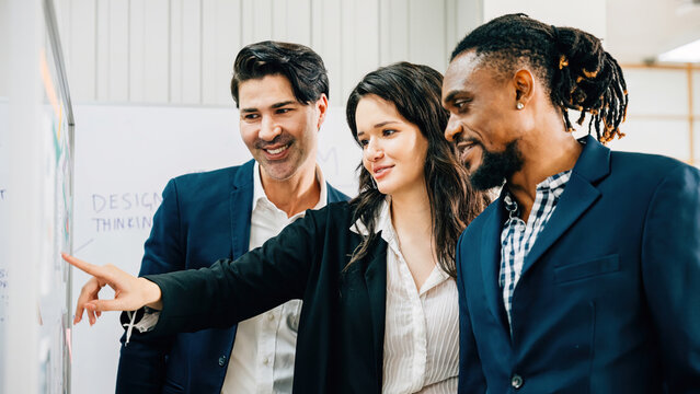 Office meeting dynamics, Business professionals employ sticky notes on a glass board for brainstorming, emphasizing teamwork, leadership, and cooperative discussions among a diverse group.