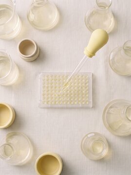 Scientific Laboratory Research Concept with Glassware and Microplate, Yellow Pipette Dropping Liquid into Wells, Minimalist Aesthetic Science Experiment Setup, Top View, Neutral Linen Background