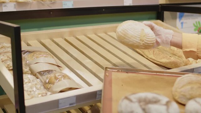 Close up view showing hands wearing protective gloves placing fresh baked baguette and artisan bread upon a wooden bakery display shelf onto a tray