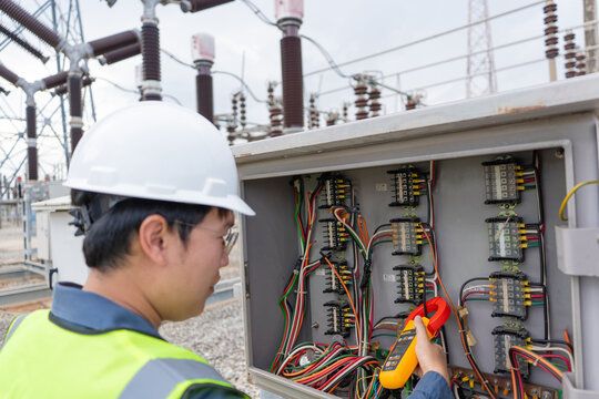 Control cabinet inspection at an outdoor substation shows an electrical technician measuring current with a clamp meter beside wiring and terminals.