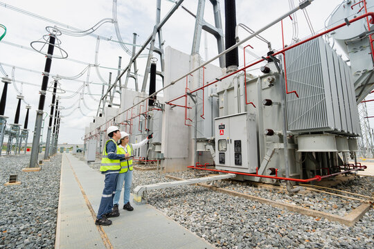 Power transformer inspection at the substation involves two engineers in hard hats holding a tablet near the control cabinet.