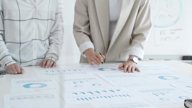 Two businesswomen examining financial data on charts and graphs during an office meeting, highlighting collaboration and strategy, close up. Business people concept