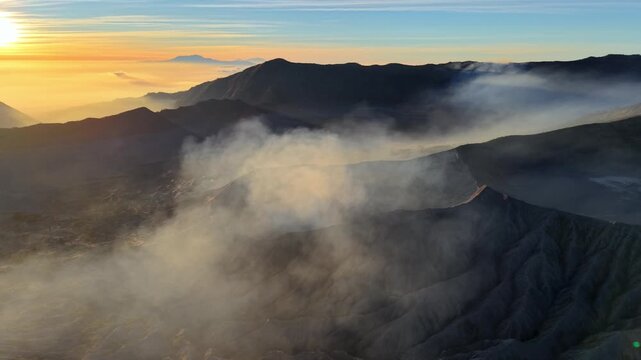 Sun rises over misty volcanic peaks. Calm, silent clouds drift through rugged terrain at Bromo Crater