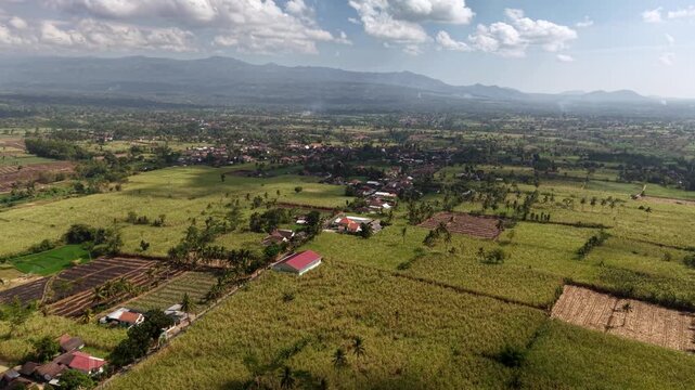 Sunlit aerial panorama of Lumajang rice paddies, villages, palm trees and distant volcanic ridges.