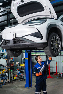 Female technician with a clipboard reaches up to check the underbody of an electric car raised on a two-post lift.