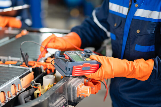 Beside an electric vehicle battery pack, gloved hands hold an insulation tester with a digital display and its red probe.