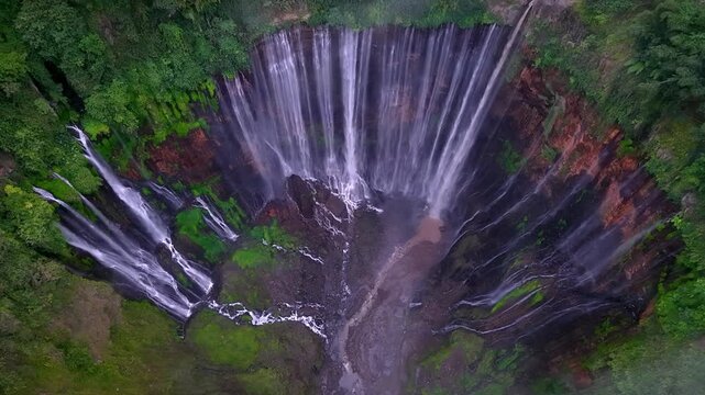 Water plunges steeply from cliffside into misty gorge. Greenery wraps rocky edges with lush at Tumpak Sewu