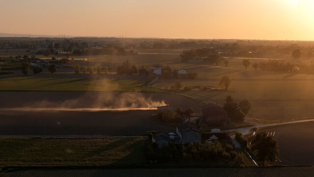 Po Valley's rural agriculture featuring vast fields and scattered farmhouses, warm sunlight creating long shadows across the land and highlighting dust plumes from ongoing cultivation work
