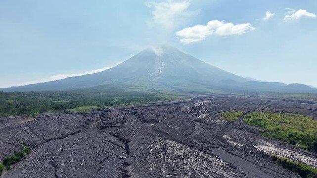 Volcano looms, ash coated earth stretches below. Sky bright, clouds drift above the jagged peaks at Desa Supiturang