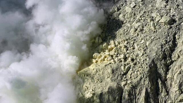 Thick white steam bursts from volcanic earth. Yellowish sulfur deposits cling to rugged rock faces at Mount Ijen
