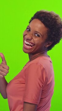 Cheerful African American woman turns around to show thumbs up before quickly switching facial expression into deep disgust against green screen. Playful lady proudly displays contrasting emotions.