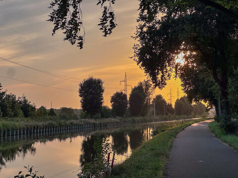 tree-lined canal under sweeping blue sky and streaked clouds, reflective water and green fields creating serene pastoral vista;