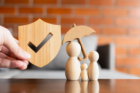 Family insurance and verified protection concept, hand holding wooden shield with checkmark near family peg dolls under umbrella on table, representing health, life, and safety coverage.