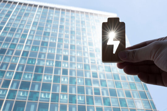 Green energy and battery storage concept, hand holding wooden battery with lightning symbol against modern office building with sun flare, representing sustainable renewable clean power.
