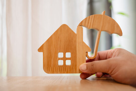 Real estate insurance and home protection concept, hand holding wooden umbrella over wooden house on desk, symbolizing property security, mortgage safeguard, and residential coverage.