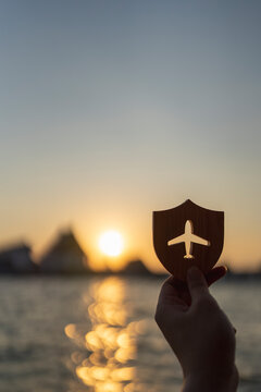 Travel insurance and flight protection concept, hand holding wooden shield with airplane silhouette at golden sunset over water, symbolizing safe journey, airline coverage, and tourist security.