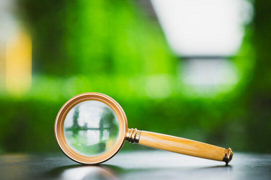 Search and discovery concept, vintage magnifying glass with wooden handle on reflective surface with blurred green nature background, representing audit, research, inspection, and analysis.
