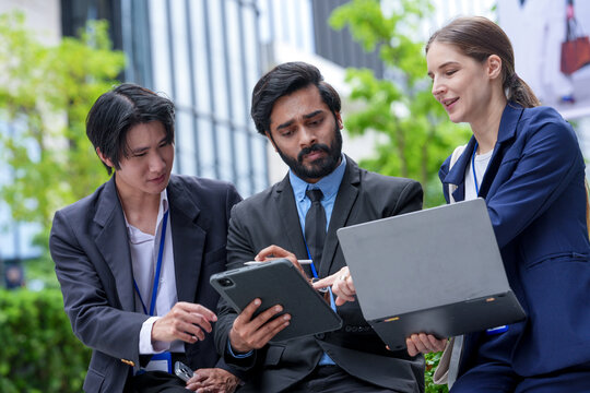 Three young business professionals wearing conference badges compare tablet notes with the laptop report in the green outdoor courtyard of a downtown business (frame 127)