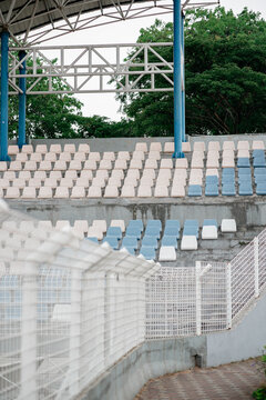 View of empty blue and white plastic seats in stadium grandstand behind a white metal fence.