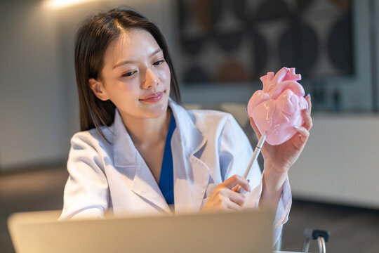 Asian female cardiology doctor holding a pink heart model for online telehealth on laptop, Medical expert explaining heart attack disease, patient education and digital healthcare consulting concepts