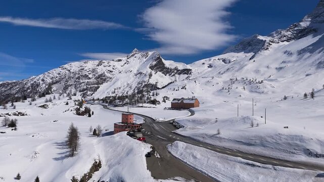 The pass road curves between deep snowfields at the Simplon Pass, Switzerland, a round stone tower and a long hospice building set against the white peaks. Aerial drone wide shot