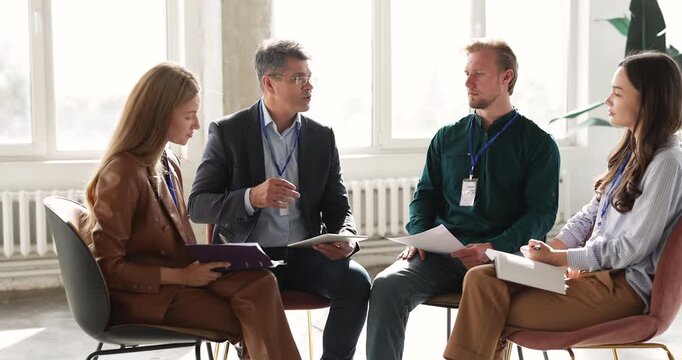 Interactive team learning workshop at corporate event with young employees sitting in circle in office taking notes after middle aged business trainer presenting strategy moderating open discussion