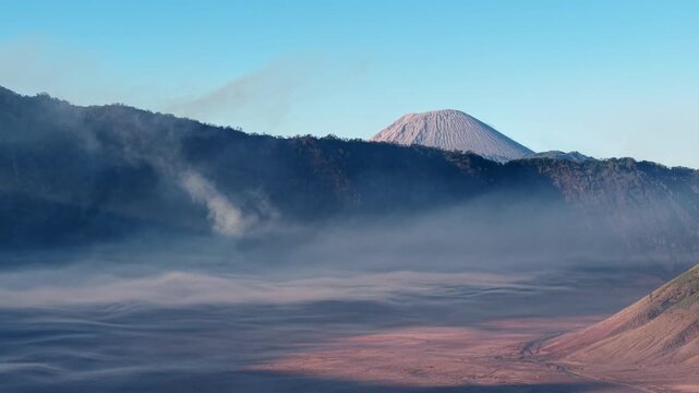 Misty dawn wraps volcanic peaks in soft blue haze. Fog drifts low across reddish earth