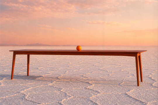 Long wooden table with single orange standing on white salt flat at sunset. Surreal minimalist scene with pastel sky and empty horizon
