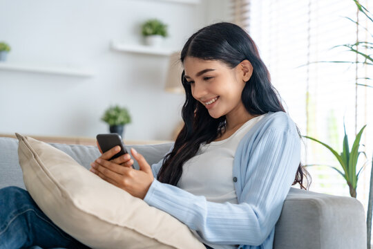 Smiling pretty young Asian woman sitting on cozy couch, using funny mobile apps in living room. Woman at Home, Doing Online Shopping, Messaging Friends, Posting on Social Media