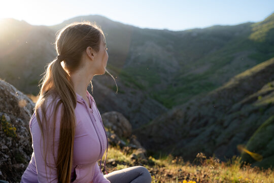  Female traveler sitting on a grassy mountain slope during golden hour, quietly observing the distant landscape. Outdoor recreation, solitude and connection with nature.