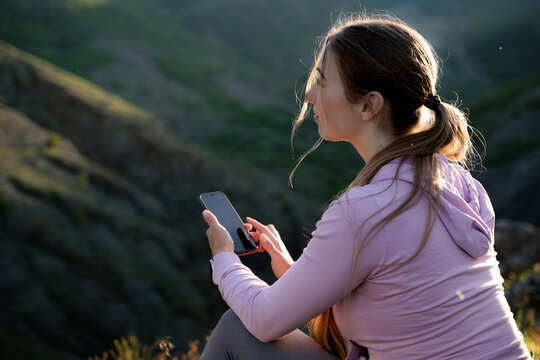 Woman in pink outdoor clothing checking her smartphone against a mountain landscape during golden hour. Digital connectivity, hiking and adventure tourism concept.