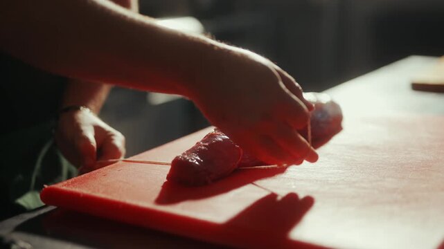 Butcher tying string around raw meat on a red cutting board