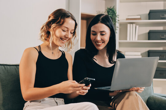 Two young female students using a laptop while sitting on a sofa at home. Student life concept with studying, teamwork, and online learning.