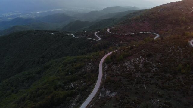 Aerial view of serpentine asphalt road cutting through dense forest and rocky terrain in high-altitude mountain region. Static elevated drone perspective captures misty valley and layered mountain.