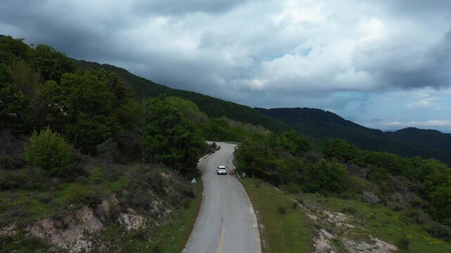 A winding mountain road cuts through green forested hillsides under cloudy skies in Livaditis, Xanthi Region, Eastern Macedonia and Thrace, Greece. Smooth aerial tracking shot follows vehicle along.