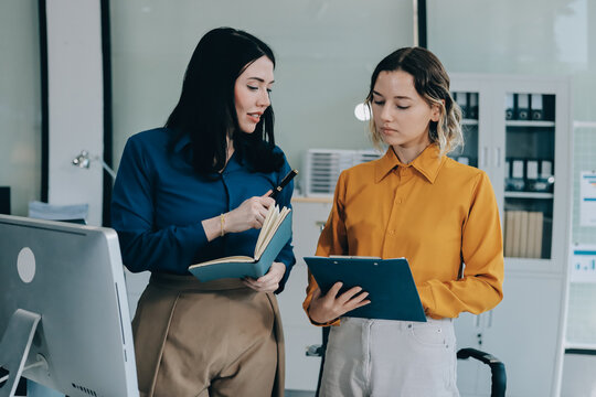 Two businesswomen discussing work using a laptop in a modern office lounge area, showing teamwork and communication
