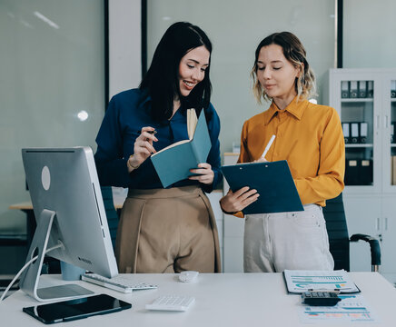 Two businesswomen discussing work using a laptop in a modern office lounge area, showing teamwork and communication