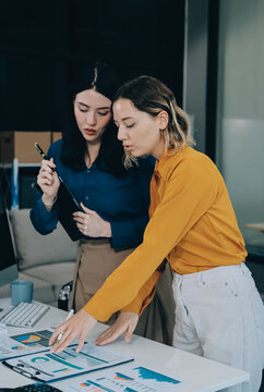 Two businesswomen discussing work using a laptop in a modern office lounge area, showing teamwork and communication