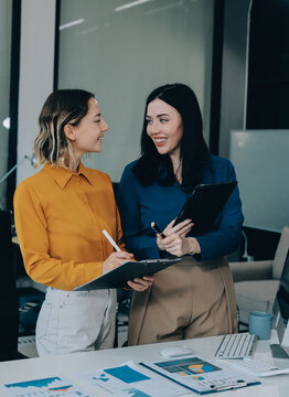 Two businesswomen discussing work using a laptop in a modern office lounge area, showing teamwork and communication