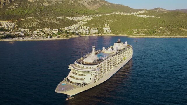 Cruise ship with a prominent bow and open upper decks lies in a calm bay off Kas Turkey with white hillside buildings beneath steep mountains in a high angle aerial view across the vessel